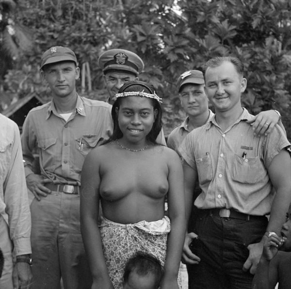 An indigenous woman poses with NATS (Naval Air Transport Service) crew on Ella Island, Tarawa Atoll, in the South Pacific. She is wearing a flowered lava lava (kilt), necklace and two headbands, one plastic and one made of a double strand of shells. Her hands are on either side of the head of a small child in front of her. The profile of a second child is on the right. The crew are wearing various uniforms. Jungle foliage is in the background.