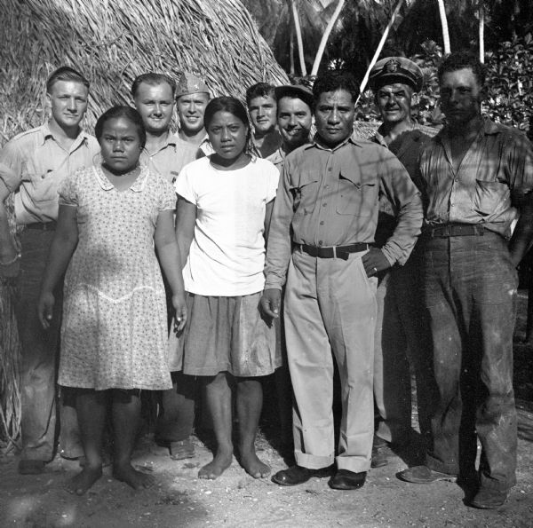 Indigenous villagers pose with Robert Doyle (7th from left) and NATS (Naval Air Transport Service) crew on Ella Island, Tarawa Atoll, in the South Pacific. The women are wearing dresses and are barefoot, and the men are wearing various uniforms or work clothes. Jungle foliage and the thatched roof of a hut are in the background.