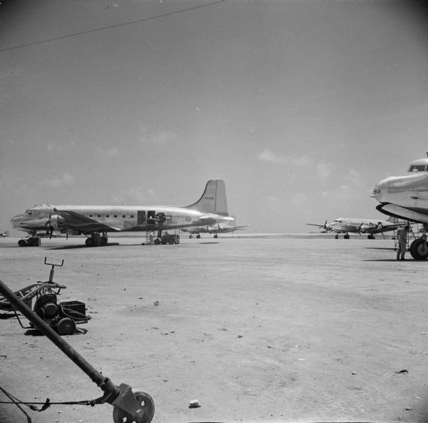 An ATC (Air Transport Command) Douglas C-54 Skymaster aircraft is being loaded by two soldiers at the air base on Kwajalein Island of the Marshall Islands in the South Pacific. Another soldier is under the nose of an airplane on the right, and beyond him is a third airplane. Machinery is in the foreground on the left.