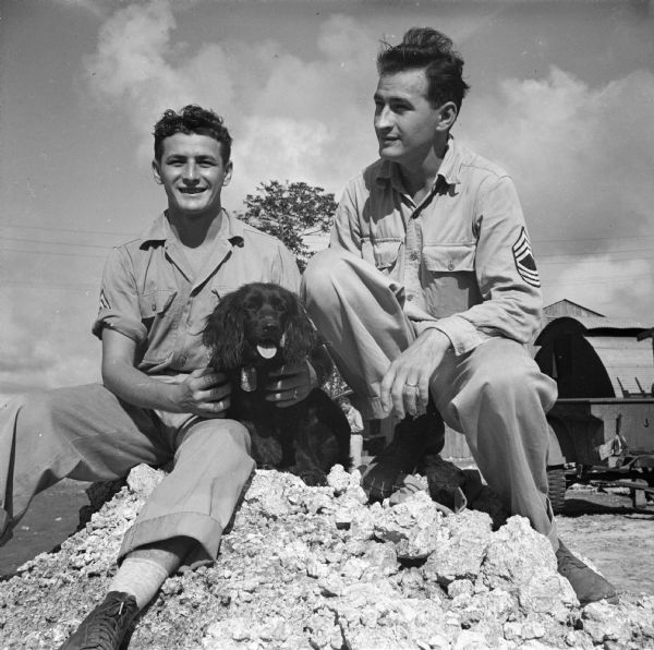 Twin soldiers from Milwaukee, Wisconsin, pose on a pile of rubble with a dog at the Army base on Guam, in the South Pacific. On the left is Corporal Robert Hugo and on the right is Master Sergeant Reinhardt (Ryan) Hugo. Although Ryan enlisted three months ahead of Robert, they work in the same quonset hut and sleep in the same pyramidal tent. Buildings and a trailer are  in the background.