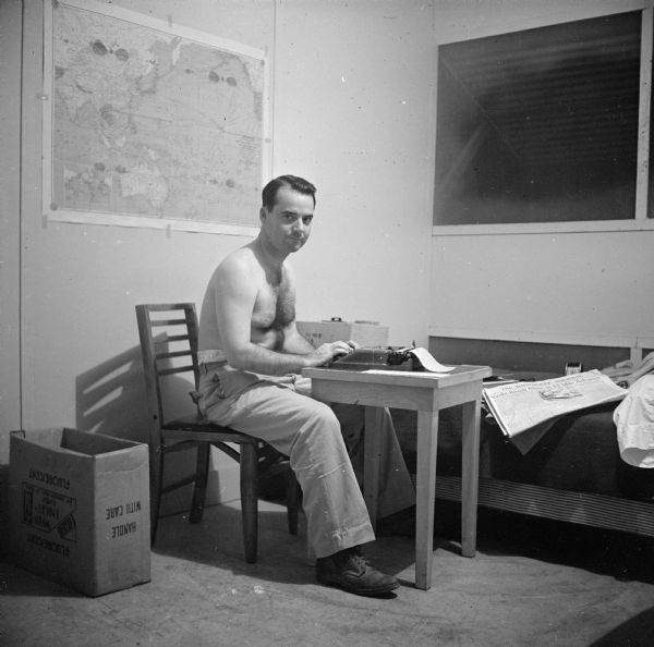 Robert Doyle (shirtles), works at the typewriter in his quarters in the BOQ (Bachelor Officer Quarters) #5, room 110, at CINCPAC (Commander in Chief, Pacific) headquarters in Guam. He has a large map of the Pacific Ocean pinned to the wall behind him. The room has simple furnishings: a table, chair and bed. A copy of the <i>Milwaukee Journal</i> is  on the bed. The shutter on his window is open. At that time, Admiral Chester W. Nimitz was the Commander in Chief, United States Pacific Fleet. On the reverse of the contact print is written, "Guam, CinCPac room."