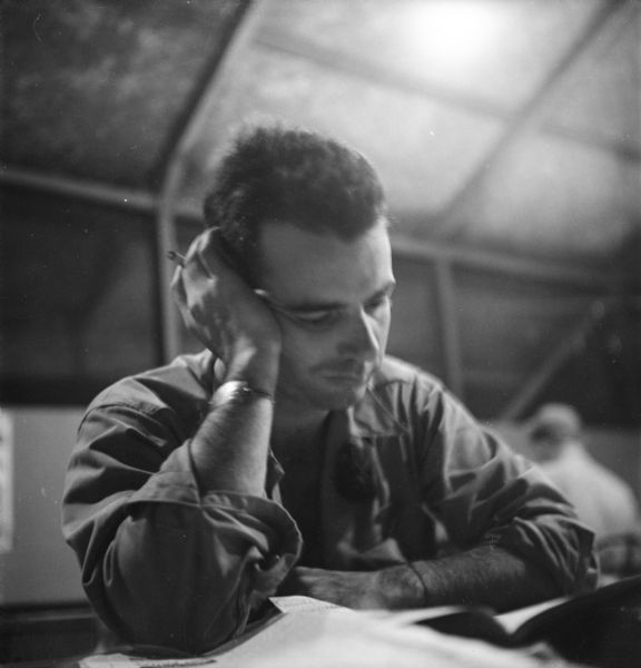 Robert Doyle as he reads and writes indoors. He is wearing a bracelet (perhaps an ID) on his right wrist, and is holding a cigarette. Another man sitting behind him.