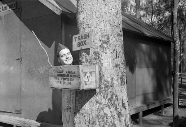 Gag photo of Robert Doyle with his head seemingly sticking out of a box. Above the box is a sign that reads: "TRASH BOX." The box and sign are attached to a tree. A building with a sign "[?]O ADMITTANCE" above the door is behind him. Trees are in the background. The location was probably Camp Cable, near Brisbane, Australia. Printed on the box is "36 TINS 16 OZ. VEGETABLE SOUP, PICK-ME-UP [???????]ENT Co1[??], SYDNEY, AUSTRALIA," and on the supporting piece of wood, "TO BASE SECTION, ARMY EXCHANGE OFF[???], BRISBANE."