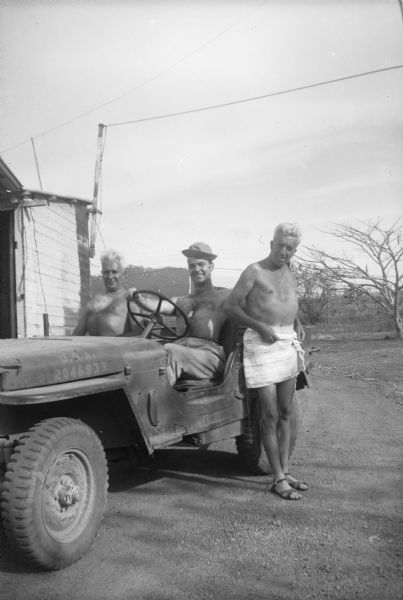 Group portrait of Robert Doyle, wearing a hat and pants, sitting at the wheel of a jeep along with a man in the passenger seat. Another man is leaning against the vehicle near Doyle. Doyle has one hand on the steering wheel and the other on the standing man's shoulder. They are going to or returning from the showers since they are shirtless, and the standing man is wearing a towel around his waist and sandals on his feet. A building is on the left and foliage and trees are in the background.