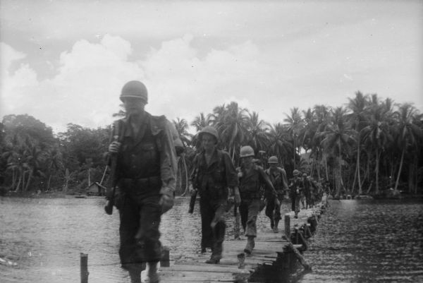 Soldiers in battle gear, holding rifles, marching in a long line across a wooden bridge. Across the water is a building on the shoreline with the jungle behind it. The location is probably in New Guinea (present day Papua New Guinea).