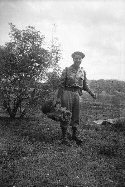 Robert Doyle poses outdoors with his gear, helmet and boots. He is standing in a field, with a tree on the left and woods in the background. A piece of sheet metal is on the ground on the right. Judging by the scrapbook kept by Doyle's mother, the location is probably "somewhere in New Guinea," (present day Papua New Guinea).