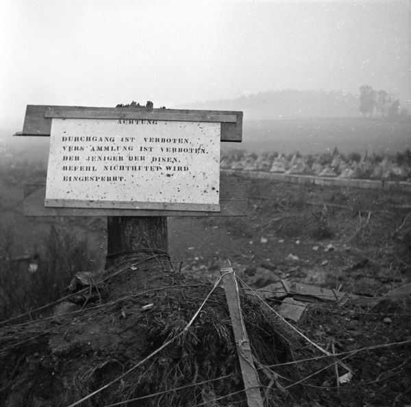 A German warning sign nailed to a tree trunk. Just beyond the sign are dragon's teeth, concrete pyramids put in to stop tanks, on the Siegfried Line, a line of defensive forts and tank defenses inside the German border. Robert Doyle took this photo on a trip from Spa, Belgium, to Aachen, Germany. He notes that it was a "miserable day, cold and misty and 'socked in'." A loose translation of the sign reads, "Caution. Passage is prohibited. Assembly is prohibited. If not obeyed, will be imprisoned."