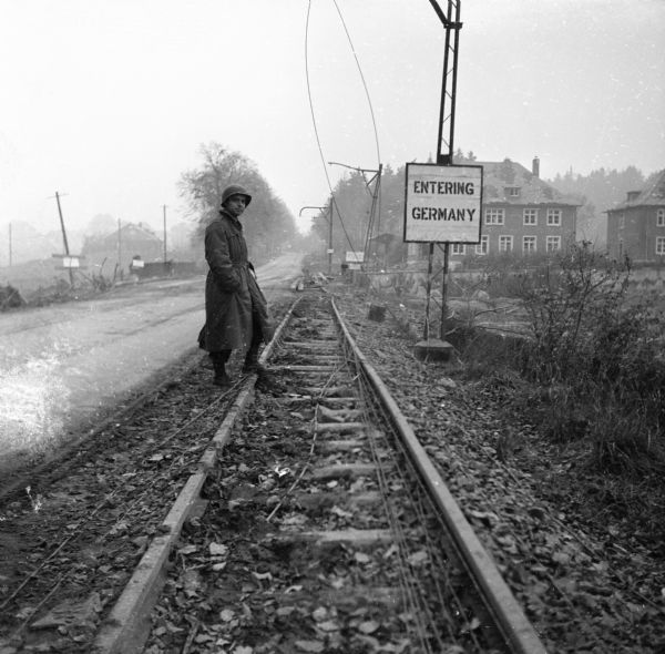 Robert Doyle stands with one foot resting on a railroad track on the border between Germany and Belgium. A sign on the right reads "Entering Germany." Behind the sign are a few buildings and trees. A road runs along the railroad tracks on the left. This image was taken on a trip between Spa, Belgium, and Aachen, Germany. Doyle notes that it was a "miserable day, cold and misty and 'socked in'." He is wearing a long coat, boots and helmet.