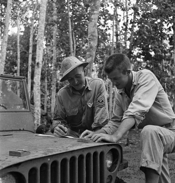 Two soldiers use the hood of a jeep as a desk as they view some papers. One man is a First Sergeant and the other has insignia on his collar but is obscured. Trees are in the background.
