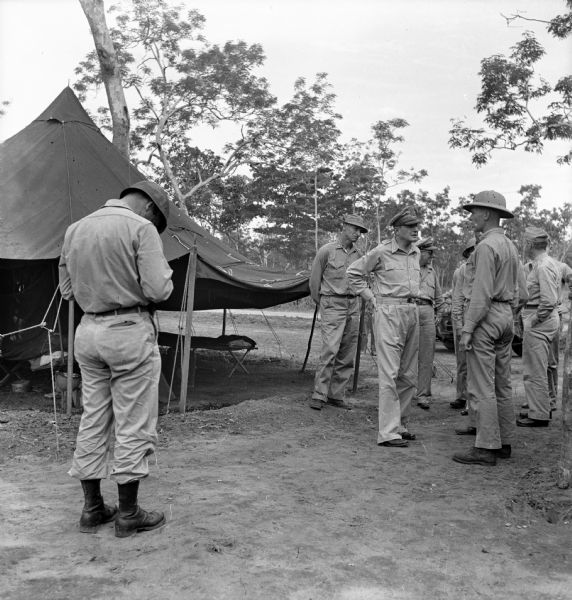 Robert Doyle takes a photo of General Douglas MacArthur as he greets a soldier during a tour on Goodenough Island, in the Solomon Sea, New Guinea (present day Papua New Guinea). Other soldiers and officers are behind MacArthur, and a tent is on the left. Trees are in the background. Handwritten on the back of the print, "Doyle snaps MacArthur."