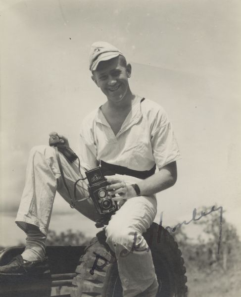 Casual portrait of photographer Sergeant Dick Hanley, smiling broadly and perched on the spare tire of a jeep, of <i>YANK</i> Army Weekly magazine. In his hands are the tools of his trade, a twin-lens reflex camera and light meter. The viewfinder cover is open and the logo for "Dick Hanley" photography appears on it. He left his civilian newsphoto beat on the streets of New York City to enlist in the Army. He ended up covering 12 different landings in the Southwest Pacific during General MacArthur's island hopping push north to Japan. He is wearing casual clothes instead of his uniform. He autographed the photo across the lower right corner.