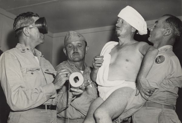 Four war correspondents pose for a gag photo. Identified (left to right) Frank Morris, <i>Collier's Magazine</i>; Harold Smith, <i>Chicago Tribune</i>; Ray Coll, Jr., <i>Honolulu Advertiser</i>; and Bob Reese, B-29 Public Relations (former <i>Chicago Tribune</i> writer). Morris is wearing a diver's mask and is apparently interviewing Coll, taking notes on a roll of toilet paper. Smith and Reese are holding up Coll, who is dressed in a towel toga and turban while smoking a cigarette. The other men are wearing uniforms.