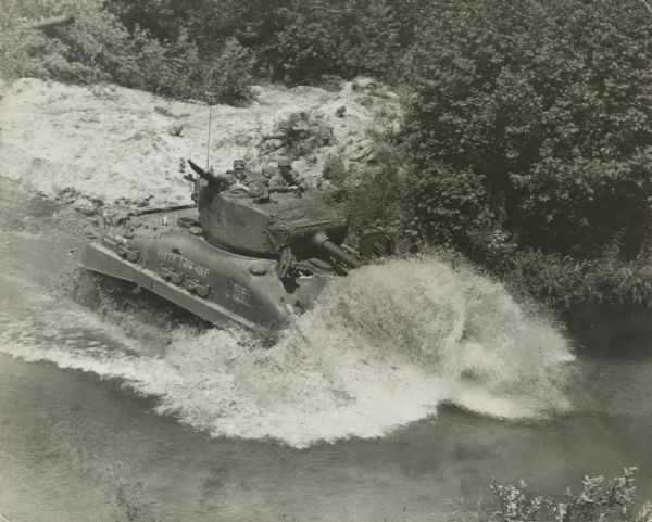 Elevated view of a tank fording a river. Two soldiers are standing in the hatches on the gun turret, with another soldier in a partially opened hatch below the turret. Foliage and trees are along the riverbank.