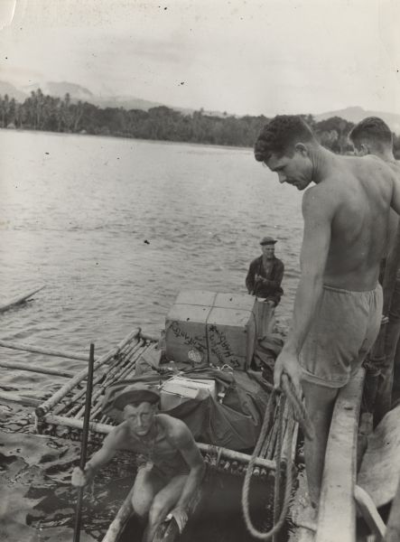 Small fishing vessels carried supplies for troops marching up the coast towards Buna, New Guinea (present day Papua New Guinea). Here soldiers lower cases over the side to an outrigger canoe for the trip to shore. Indigenous crews helped load and unload supplies. The tree-covered shore and hills are in the background. Photograph taken by Sergeant Leland Score of Menomonie, Wisconsin and selected for publication by Robert Doyle. Sergeant Score was a clerk at division headquarters and carried his camera with him through the Papuan campaign. He was taking the photographs to be used by the division for an official history after the war.