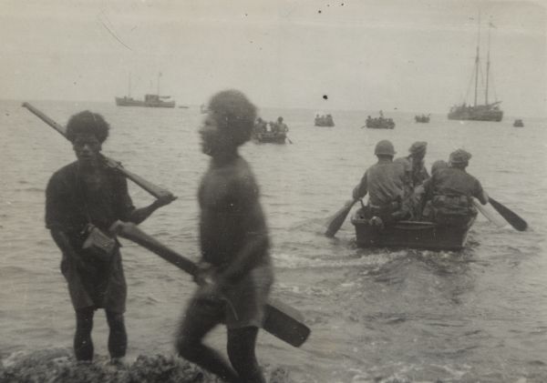 Indigenous men help 32nd Division soldiers from units, originally from Wisconsin, who paddle in collapsible canvas boats to small coastal vessels. The man with the rifle (left) is a "police boy," who speaks some English and bosses the others. Text in the <i>Milwaukee Journal</i> describing this group of images reads: "War Correspondent's Album. These pictures, made in New Guinea last November, December and January by Robert J. Doyle, Milwaukee Journal war correspondent, have just been received, approved for publication by the photo news board of the bureau of public relations of the war department."