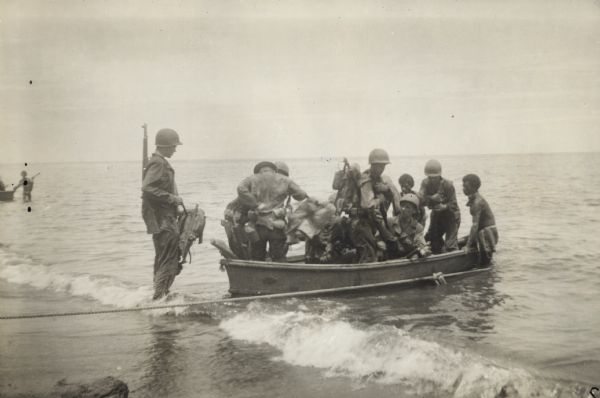 Soldiers land on the beach in New Guinea (present day Papua New Guinea). They use collapsible canvas boats to ferry men and equipment from small coastal vessels. Indigenous men help paddle the boats.