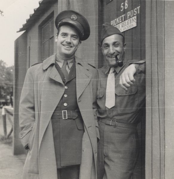 Robert Doyle, and Corporal Sam Oxman of the Public Relations Office, smile and pose outdoors in front of a door at Earls Colne Airfield, Essex, England. Corporal Oxman is from Philadelphia, Pennsylvania. The information on the door reads "58 Picket Post, Public Relations, [?]'s Hacienda." Corporal Oxman is in uniform and is smoking a pipe. Doyle wears a uniform with an overcoat and a US War Correspondent hat.
