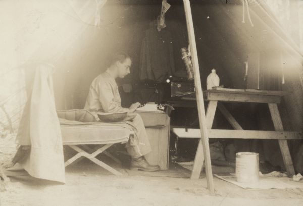 Robert Doyle types stories about the Red Arrow Division in his tent while seated on his cot. The tent sides are tied open. A crate serves as a typewriter stand and a canteen is on a picnic-style table on the right. The can beneath the table has a label that reads "Defense Pure Coffee." A flashlight is tied to the tent pole and other equipment and clothing are behind Doyle. On the reverse of the print is written: "Australia," so the location may be Camp Cable, south of Brisbane.