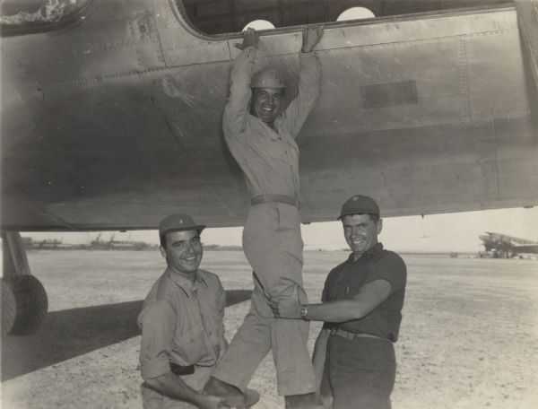From the caption that accompanied the image: "Fellow workers in civilian life, these Milwaukeeans met recently on Kwajalein atoll in the Marshall islands. They are (from left) Robert J. Doyle, The Journal's staff war correspondent; Lieutenant Louis Olszyk, on leave from The Journal news staff, now officer in charge of the 4th marine air wing news service at Kwajalein, and Ensign Ray Hansen, also on leave from The Journal news staff, who works in the naval air transport service office at Kwajalein." They are all smiling, and Doyle and Hansen hold Olszyk's legs as he reaches his arms up to hold onto the bottom of an open airplane door. Another airplane is in the far background on the right.