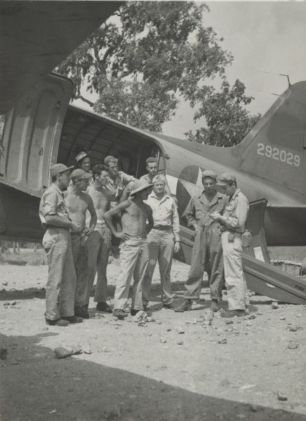 Robert Doyle interviews a group of nine soldiers at an airfield. They are standing in front of an airplane (tentatively identified as a Douglas C-47 Skytrain passenger and cargo aircraft) with the side door open and the loading ramp down. From the various uniforms and hats, they may be from several branches of the service. Some of the men are shirtless. Trees are in the background. On the reverse of the print is written, "From Myron H. Davis." Davis was a photographer for <i>Life Magazine</i>.