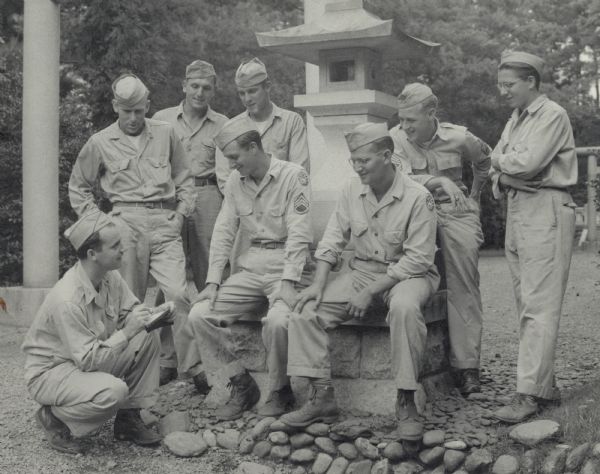 A shinto shrine near Tokyo serves as the starting point of a Wisconsin story as Robert J. Doyle (lower left), correspondent of the Journal, takes the names of a group of state soldiers. All members of the 5th Air Force headquarters, they are (left to right) Private First Class Joseph Borkenhagen, Private First Class Alvin Schultz and Private First Class Arthur Grudnowski, all of Milwaukee; Staff Sergeant Harry Kallish of Wisconsin Rapids; Sergeant Ervin Erlein of Milwaukee; Technical Sergeant Robert Sheldon of LaCrosse; and Sergeant Micheal Pokos of West Allis. The above information was taken from the caption that ran with the image in the <i>Milwaukee Journal</i>. The shrine was located near the 5th Air Force Headquarters near Tokyo, Japan.