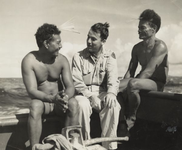 Micronesian men, who put thatched roofs on buildings in the western Caroline Islands for United States forces, sit chatting with Robert J. Doyle, The Journal's staff war correspondent. They are sitting on the gunwales of a harbor tug with the ocean behind them, and are located near Ulithi, Caroline Islands, in the Western Pacific. According to Doyle, the men wear bamboo combs in their hair as a sign that they are bachelors, looking for a girl. If they hand the comb to a girl and she hands it back, it's no soap. If she keeps it, there's a wedding. The above information was taken from the caption that ran with the image in the <i>Milwaukee Journal</i>. Doyle is sitting between the bachelors, and a wind is blowing their hair. Doyle is in uniform and the men are wearing lap-laps (loincloths).