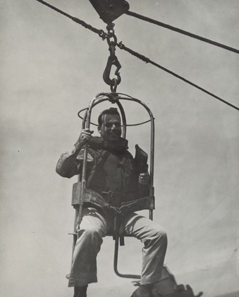 A smiling Robert Doyle is transferred between two ships using a manila highline in a transfer-at-sea chair, also called a boatswain's chair. He is wearing a life preserver and carrying a book.