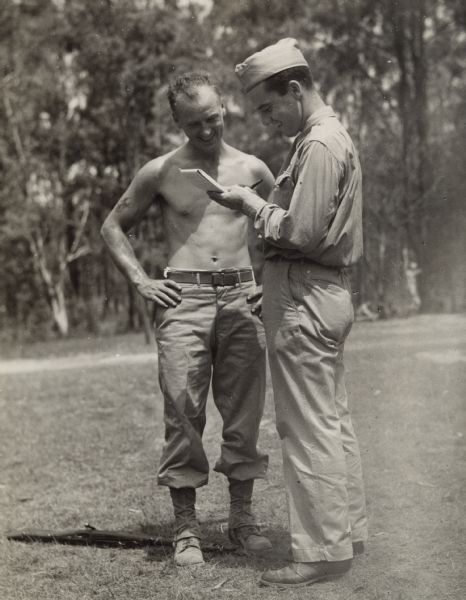 Robert Doyle interviews Private Floyd Flayter, (shirtless) of Milwaukee, Wisconsin at a training ground somewhere in Australia. A firearm is laying on the ground under his feet. Private Flayter had this to say: "Tell the folks in Milwaukee that slamming around in the unarmed combat training course being given American forces in Australia is a bit different from working in a shoe factory." A field and trees are in the background. When the image was published in the <i>Milwaukee Journal</i>, the background was blacked out.