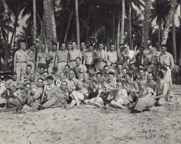A group of war correspondents, soldiers and officers pose outdoors on the sand in front of palm trees for a portrait in Guam. Many are holding a beverage and plate. In the lower right corner is written, "Guam, April 1945." All identified people have a number written on them in ink. Their corresponding names were typewritten on a separate piece of paper. Listed are 1. Wally Soderholm (<i>Buffalo News</i>); 2. Bob McCormick (Radio); 3. Elmonte Waite (Associated Press); 4. Robert Doyle (<i>Milwaukee Journal</i>), (in list named Dopey Doyle); 5. Carl Mydans (<i>Life</i>); 6. Tom Stowe (Red Cross); 7. Phil Heisler (<i>Baltimore Sun</i>); 8. Walter Davenport (<i>Colliers</i>); 9. Chu Chiping (Ta Kung Pao, Chungking); 10. Gene Rider (Columbia Broadcasting System); 11. Murlin Spenser (Associated Press); 12. David Brown (Reuters); 13. Larry Tighe (Blue Network); 14. Joe Bors (International News Service); 15. Graham Stanford (<i>London Daily Mail</i>); 16. Frank Tremaine (United Press); 17. John Brennan (Aussie Papers); 18. Captain Fitz-Hugh Lee (PRO); 19. Leif Erickson (Associated Press); 20. Captain Min Miller (PRO), H.B.; 21. Shelley Mydans (<i>Life</i>); 22. Emmett Crozier (<i>New York Herald-Tribune</i>); 23. Palmer Hoyt (<i>Portland Oregonian</i>); 24. Bruce Rae (<i>New York Times</i>); 25. Barbara Finch (Reuters); 26. Jack Mahon (Mutual); 27. Lyle Schumacher (United Press); 28. Henry Keyes (<i>London Express</i>).