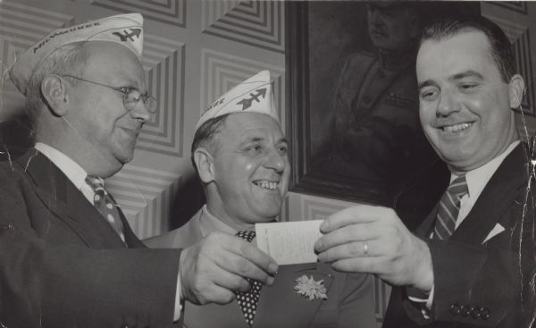 For contributing to the morale of state troops in the South Pacific by his work as a war correspondent, Robert Doyle (right), a <i>Milwaukee Journal</i> reporter, was given a life membership card in the 32nd Division Veteran association at a meeting Tuesday at the Red Arrow Club. The presentation was made by Joseph Hrdlick (left), national secretary, and Eugene Stephan, president of the Milwaukee Red Arrow club. The above information was taken from the caption that ran with the image in the <i>Milwaukee Journal</i>. All three men are wearing suits, and the presenters are wearing their Red Arrow side caps (garrison caps).