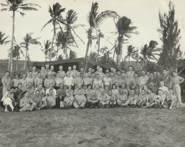 Group portrait of Fleet Admiral Chester W. Nimitz and his staff at CinCPac/CinCPOA Advanced Headquarters on Guam. Admiral Nimitz is 9th from the left in the second row. Robert Doyle (no shirt) is standing in the third row to the right of a woman sitting next to Nimitz. The event appears to be an outdoor concert. Several men on both ends of the group are holding musical instruments, and many people are holding beverages. Most of the staff are wearing uniforms, but several are wearing bathing suits. A black dog is sitting 5th from the left in the first row. In the background is a tent and palm trees.