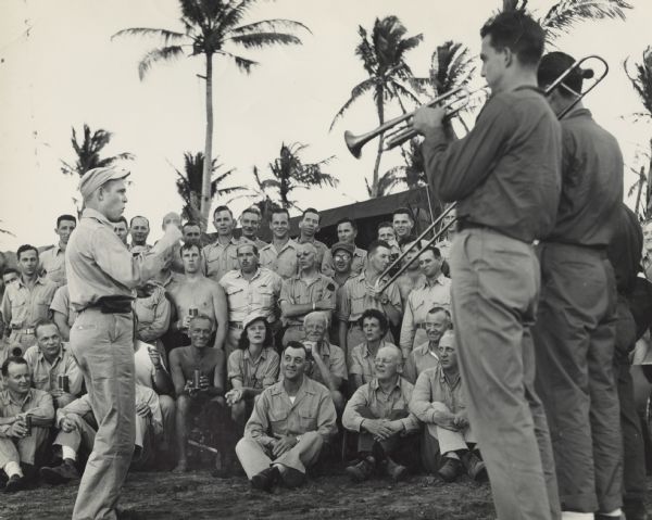 An outdoor concert for Fleet Admiral Chester W. Nimitz and his staff at CinCPac/CinCPOA Advanced Headquarters on Guam. Admiral Nimitz is 3rd from the right in the second row. Robert Doyle is 2nd from the right in the third row, behind the trombone slide. The conductor is standing on the left and the band is on the right. Most of the staff are wearing uniforms, but several are wearing bathing suits. Many members of the audience are holding beverages in cans. A black dog is sitting in the first row. In the background is a tent and palm trees.