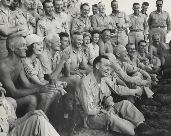 The audience laughs or sings at an outdoor concert for Fleet Admiral Chester W. Nimitz and his staff at CinCPac/CinCPOA Advanced Headquarters on Guam. Admiral Nimitz is 3rd from the left in the second row. Robert Doyle is 3rd from the left at the top (No shirt and only the bottom of his face appears). Most of the staff are wearing uniforms, but several are wearing bathing suits. Many members of the audience are holding beverages in cans. A black dog is sitting in the first row.