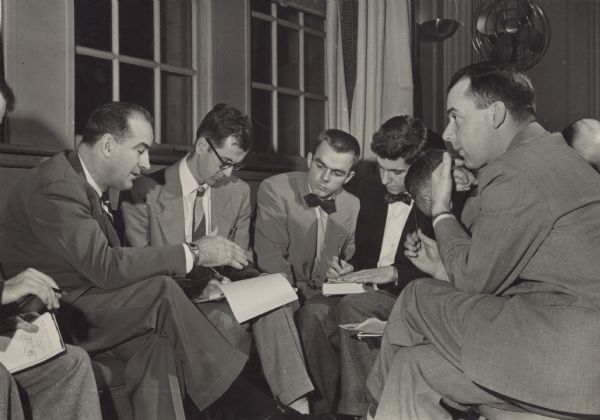 Press interview at the Memorial Union, University of Wisconsin-Madison. Identified (left to right) are the United States Senator from Wisconsin, Joseph McCarthy; Dave Lippert, <i>Milwaukee Sentinel</i>; Jerry Schecter, student reporter, <i>Daily Cardinal</i> and Robert Doyle, <i>Milwaukee Journal</i>. All the men are wearing suits and most have notebooks on their laps.