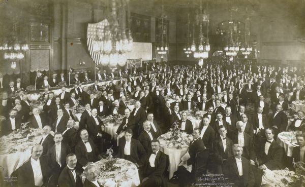 Elevated view of tables of seated diners at a banquet tendered to Hon. William Howard Taft by the Merchants and Manufacturers Association of Milwaukee at the Hotel Pfister. Taft appears to be seated directly beneath the shield at a long table along the back wall on the left.