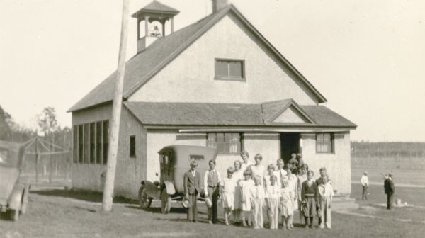 Pupils and their teacher (rear) pose in front of their school house. Two cars are parked in the school yard. A man and other pupils stand near a water pump on the right; there is a merry-go-round behind the school on the left. The school bell can be seen in its tower.