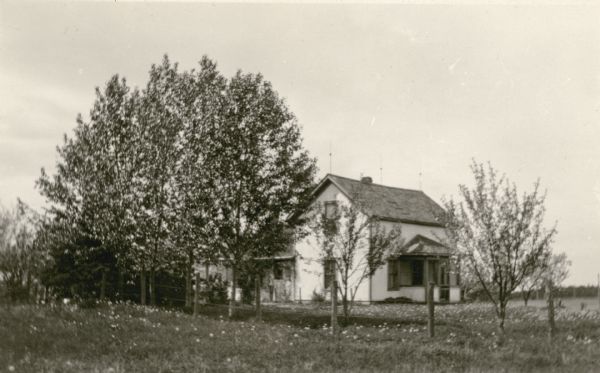 A one and a half story wood frame house stands behind a wire fence.  Wildflowers bloom in the yard and there are several trees near the house. Lightning rods are on the ridge of the roof. This was the home of Mrs. Inez Lindell Bystrom, teacher at the Miles School.