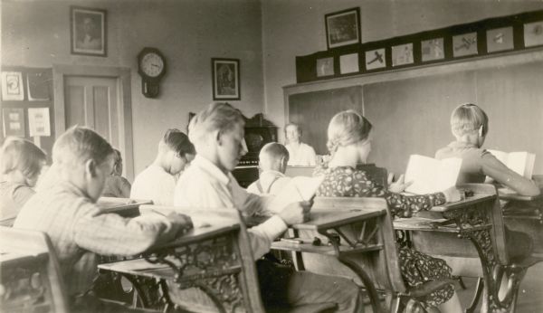 Students of the Miles School, Bayfield County District No. 2, read at their desks supervised by their teacher, Mrs. Inez Lindell Bystrom. There is a parlor organ at the front of the room and a clock hangs on the wall near a portrait of George Washington.