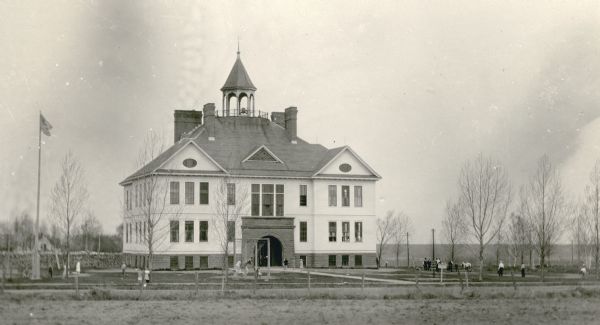 Children play on swings in front of the imposing Port Wing Consolidated School. It is two stories tall, with wood frame construction, a tall sandstone foundation and four large chimneys. The arched front entry is also stone. There is a bell tower with a finial. Cord wood is stacked to the left of the school, behind a large flag pole. This was the first consolidated school in Wisconsin.