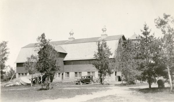 An automobile is parked near the Melvin Larson barn in Bayfield County. The large barn has a concrete block foundation, gambrel roof, and three large ventilators, each with a weather vane. There is a silo on the right.