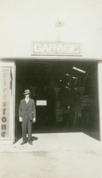 Raymond Alpin, president of the Potter School District No. 3 PTA stands in the doorway of his garage. There is a large Firestone advertisement on the left. A sign for Eveready Prestone Anti-Freeze is visible in the garage.