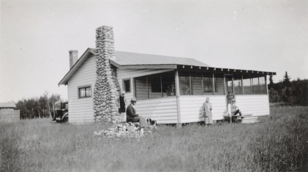 Four people and a dog pose in front of a small cottage with a large fieldstone fireplace. They are, from left: Armin Duchow and his father George, Potter, Wisconsin, and Mrs. and Mr. Alb. Hernke, Hilbert. A car is parked behind the cottage on the left. There is a child standing inside the screen door, and two other people are on the porch. The photograph was submitted as an example of "Where Pupils Travel" to the Wisconsin Rural Schools Survey. Armin Duchow was a student at Potter School, District No. 3, Town of Rantoul.