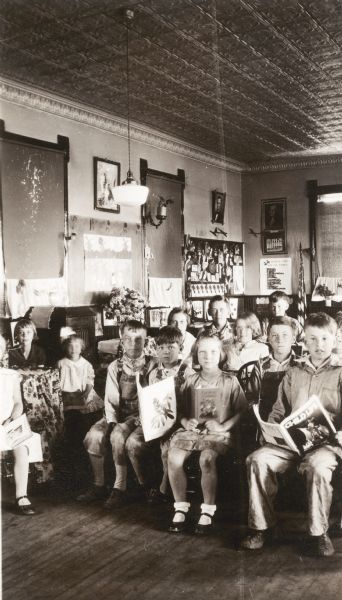 Students holding books and magazines pose seated in chairs in the Prairie School. Model airplanes hang near the portraits of Abraham Lincoln and George Washington in the back corner. The room has a pressed tin ceiling.