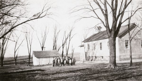Students pose in front of a small outbuilding near the Prairie School. The school has a stucco exterior and a basement. There is a bell tower on the roof.