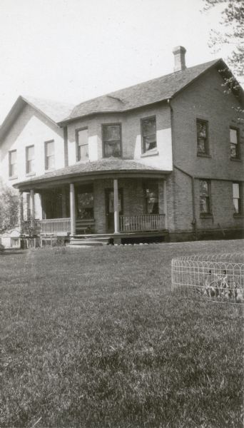 A two-story brick house, identified as the home of Prairie School pupil Marian Hintz, stands on a well tended lawn. There is a large front bay and curved porch with pillars. A low wire fence protects a flower bed.