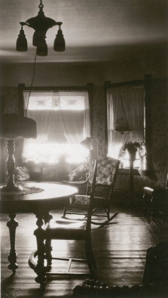 Sunlight from the front bay window illuminates the parlor of the Vickers house, identified as the home of Marian Hintz, a pupil at the Prairie School. A cord runs from the ceiling fixture to an electric table lamp. There is a potted plant in the window.