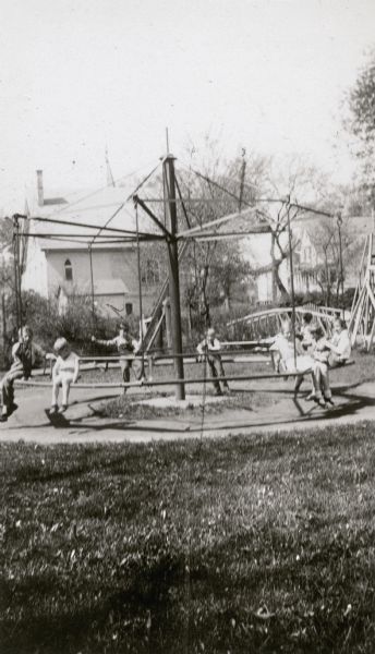 Children pose on a merry-go-round (carousel) in the Edgerton Public Park. The rear wall of a church forms the background, and there appears to be a bridge on the right.