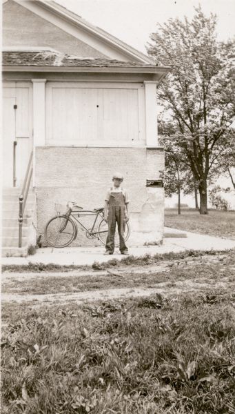 Robert Hippe, 12, in cap and bib overalls, poses in front of the Prairie School. His bicycle rests against the porch of the school. A note on the reverse of the photograph reports he "invented an airplane. Works on radios and boy's toys."