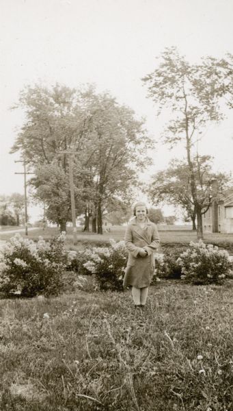 Harriet Sundbakken, 13, a student at Prairie School, poses near some flowering bushes. The school building is in the background. A note on the reverse of the photograph reports that she "received second prize for first-year sewing at the Stoughton festival."