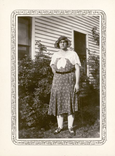Mrs. Mabel Crosby, the teacher at Peck School, poses at the side of a building. She is wearing eyeglasses, and a summer dress and hat.